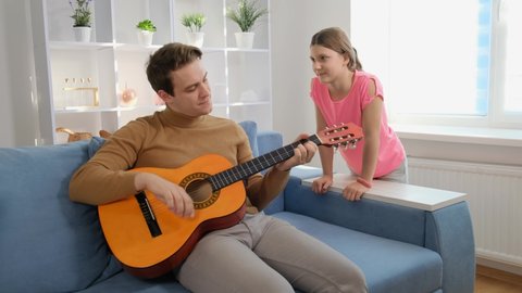 A young guy sits at home on the couch and plays guitar next to his younger sister.