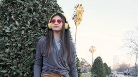 Asian woman wearing wireless headphones enjoying listening to music and dancing outdoors in the city street.