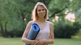 Young woman holding mat and smiling for camera while standing in green park outdoors spbi. 4k video Portrait of beautiful european female looks with toothy happy smile and holds training rubber in