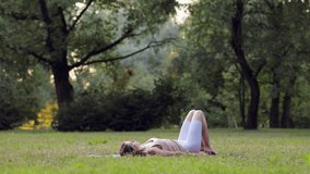 Young beautiful woman doing yoga exercise and lying on green grass outdoors spbi. 4k video Caucasian female does asana and stretches body, exercises slowly and lies on mat on background of trees in