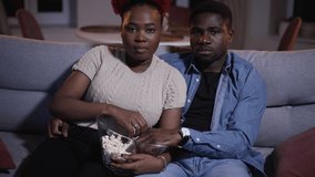 Two family people eating popcorn and having good time together on couch in home room spbas. 4k video Closeup view of beautiful african woman, man feed each other and look with smiles, hug tenderly and
