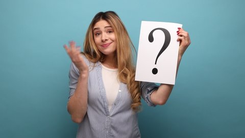 Portrait of confused thoughtful woman looking at camera, holding paper with question mark, thinks about tasks, wearing striped shirt. Indoor studio shot isolated on blue background.