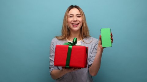 Bonus for mobile user. Portrait of happy woman holding gift box and cell phone with empty display for online shopping advertising, wearing striped shirt. Indoor studio shot isolated on blue background