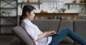 Asian business woman video chatting with laptop on couch at home. Female student talking and looking at laptop screen while resting on couch at home living room. Webcam webinar concept