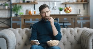 Young bearded man is sitting on the couch at home and watching TV. Bored millennial male uses the remote to switch channels in search of interesting video content and eats popcorn