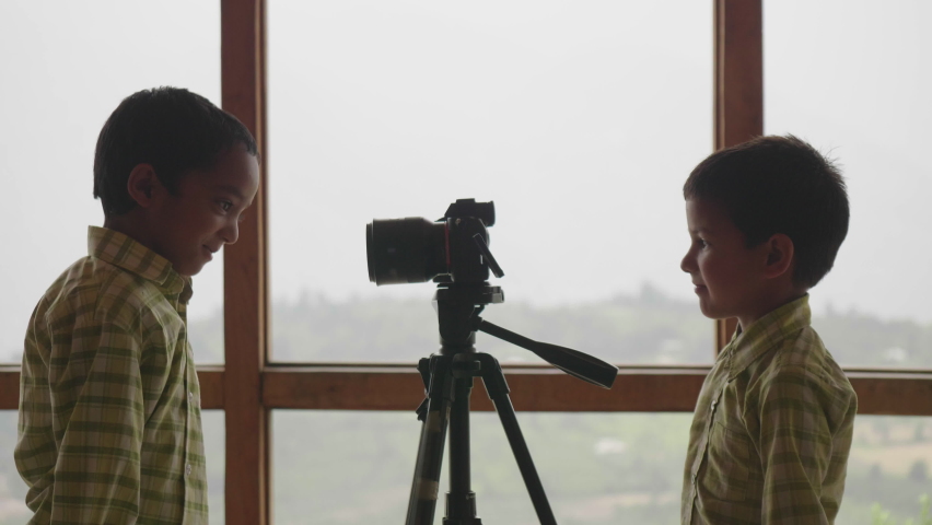 Two young cute Indian male school kids are playing with or making funny faces in front of a digital photography camera or DSLR with a background of misty mountainous valley out of window. | Shutterstock HD Video #1087418726