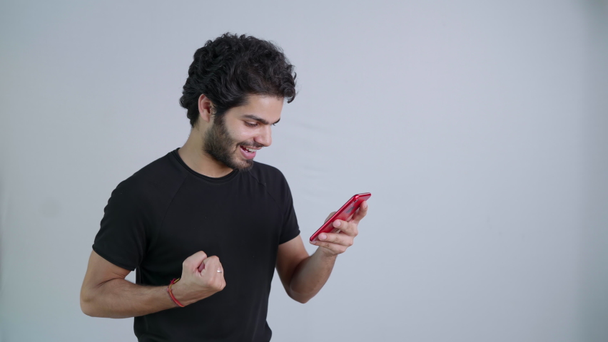 Young Indian Asian happy smiling attractive man in a black t-shirt is celebrating after a win or a victory while watching an online match or playing game on a mobile phone isolated on white background