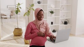 Happy Pregnant African American Woman Using Laptop Browsing Internet Wearing Hijab Sitting At Home. Freelance And Distance Job During Pregnancy. Childbirth And Modern Lifestyle. Slow Motion