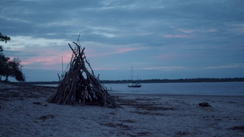 A teepee made of various sized sticks sits on the peaceful beach as the retreating sun spills pastel colours of pink and blue into the clouds, ocean and water whilst a boat floats along the shoreline