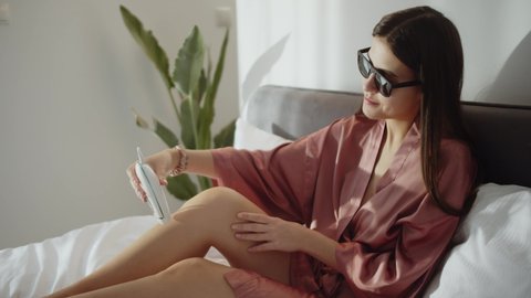 Young Beautiful Woman Shaving Leg In Pink Bathrobe In Bedroom In House Care For The Skin And Preparing To Date Or Wedding  Woman Using Laser Epilator For Hair Removal Procedure Beauty Concept