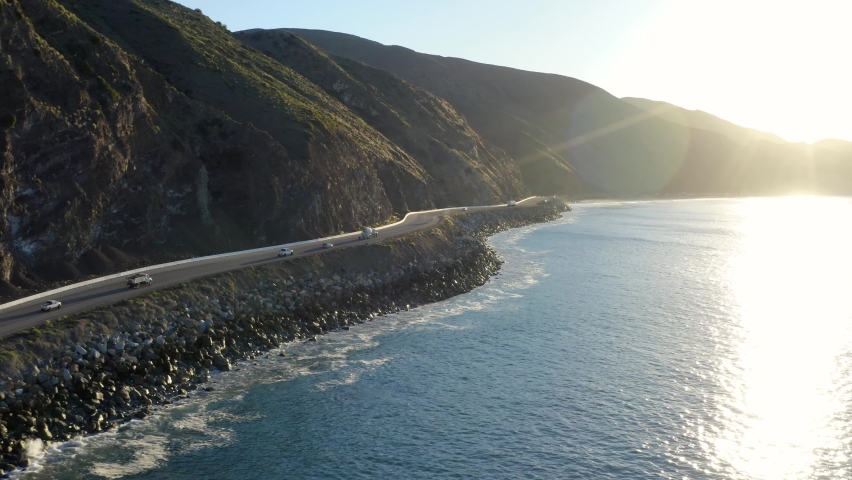 On the open road, ready for a road trip. Aerial view of a road running between a mountain and the ocean. Top view of a coastline road and beach resorts at sunset. Malibu Beach coastline in California Royalty-Free Stock Footage #1087445450