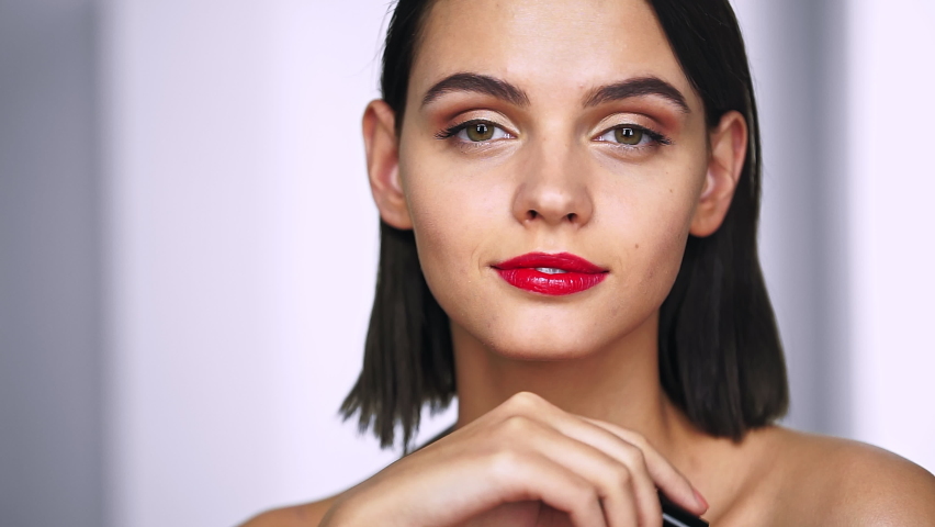 Beautiful girl painting her lips with bright red lipstick, then draws heart on the mirror and winks. Beauty