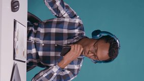 Vertical video: African american man recording and singing song on smartphone with audio headset. Happy adult listening to mp3 music and doing karaoke session, using mobile phone as microphone.