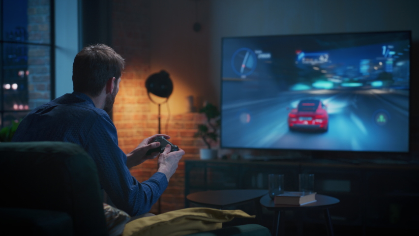 Young Man Spending Time at Home, Sitting on a Couch in Stylish Loft Apartment and Playing Arcade Car Video Games on Console. Male Using Controller to Play Street Racing Drift Simulator. | Shutterstock HD Video #1087466114