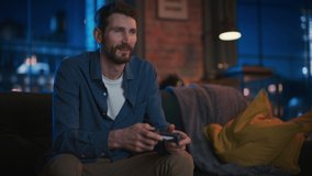 Portrait of Young Handsome Man Spending Time at Home, Sitting on a Couch in Stylish Loft Apartment and Playing Video Games on Console. Successful Male Using Controller to Play Action Game and Win.