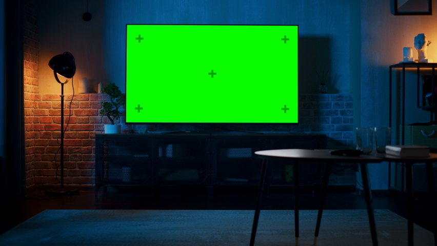 Stylish Loft Apartment Interior with TV Set with Green Screen Mock Up Display Standing on Television Stand. Empty Living Room at Home with Chroma Key Placeholder on Monitor. Zoom Out Evening Shot. Royalty-Free Stock Footage #1087466222