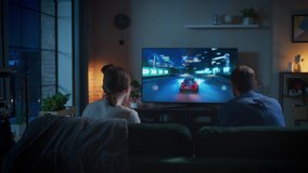 Young Couple Spending Time at Home, Sitting on a Couch in Loft Apartment and Playing Arcade Car Video Games on Console. Boyfriend Playing Drift Simulator while His Girlfriend is Rooting and Cheering.