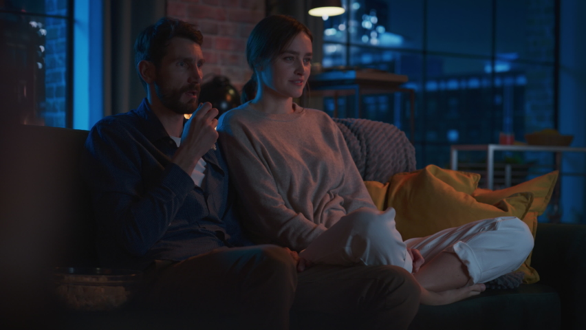 Portrait of Beautiful Couple Spending Time at Home, Sitting on a Couch, Watching Scary TV Show in Their Stylish Loft Apartment. Man Puts Hand on Female Knee, While Streaming Thriller Movie. Royalty-Free Stock Footage #1087466315
