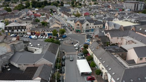 Aerial ascending footage of traffic on roundabout in town centre. Cars parked along road in front of buildings around square. Ennis, Ireland