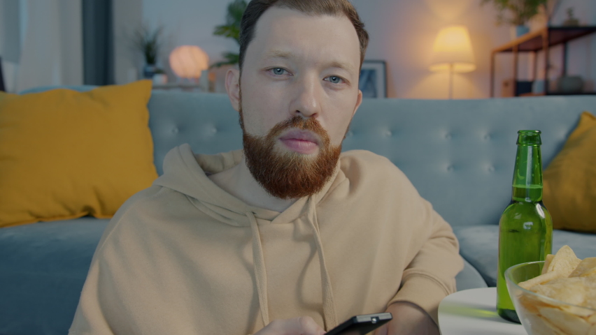 Unhappy man choosing channel on TV using remote control and looking at camera with disappointment at home at night. People and television concept.