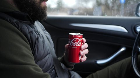 WROCLAW, POLAND - FEB 22, 2022: Handsome adult bearded man driving a car hold drinking a coca cola can sugary carbonated beverage