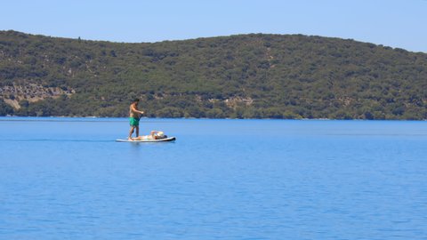 Sainte-Croix-du-Verdon, France - August 2021 : Paddleboarding tourist carrying his wife lying on the board on the Lake of Sainte-Croix on a summer day on the Verdon valley