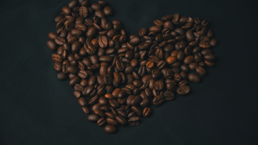 Coffee beans lie in the form of a heart, their female hand cleans them. A young girl interferes with brown fried coffee beans on a black surface. Fried coffee.