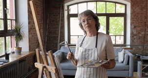 Smiling elderly female artist stand at personal creative studio pose at workplace with brush and paints on palette. Skilled aged woman professional art teacher look at camera start video drawing class