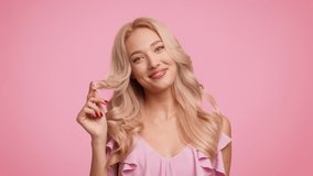 Attractive Blonde Woman Playing With Curled Hair Smiling To Camera Playfully Posing Standing Over Pink Studio Background. Haircare And Hairstyle, Female Beauty Concept. Slow Motion