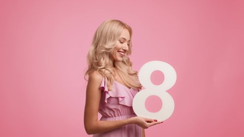 Women's Day. Happy Blonde Lady Holding Number Eight And Winking Eye, Celebrating International Woman's Holiday And Smiling To Camera Posing Standing Over Pink Studio Background