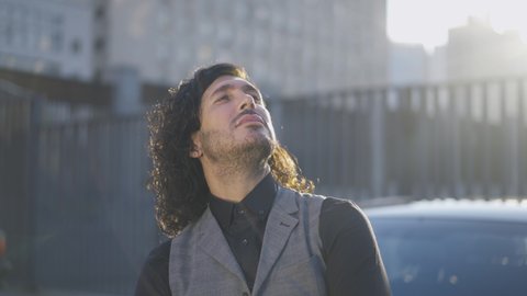 Young Caucasian man with green eyes and long hair looking up at sunshine standing in urban city. Portrait of confident handsome businessman in elegant shirt and waistcoat outdoors. Slow motion