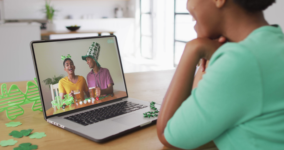 Smiling african american male friends with beer wearing clover shape items on video call on laptop. irish tradition and st patrick