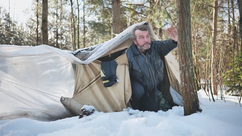 A homeless man climbs in and out of a tent in the woods in winter.