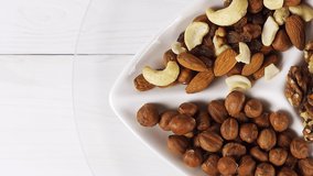 Various nuts close-up on a white plate on a white wooden table. View from above. 4k raw video with smooth camera movement.