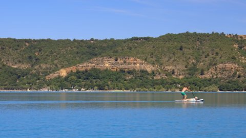 Sainte-Croix-du-Verdon, France - August 2021 : Paddleboarding tourist carrying his wife lying on the board on the Lake of Sainte-Croix on a summer day on the Verdon valley
