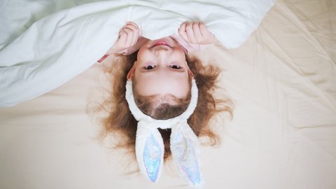 Funny surprised little girl covering half of face with white blanket. Baby girl peeking from duvet, feels embarrassed. Easter holiday. A cute little girl with bunny ears.