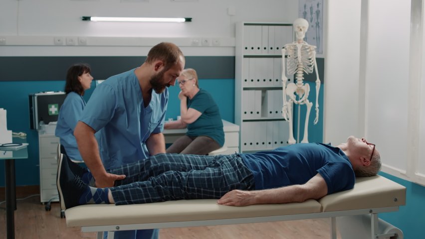 Medical assistant supporting old man in doing physical exercise to stretch legs muscles and crack bones. Specialist using alternative medicine procedure to increase mobility for patient. Royalty-Free Stock Footage #1087518227
