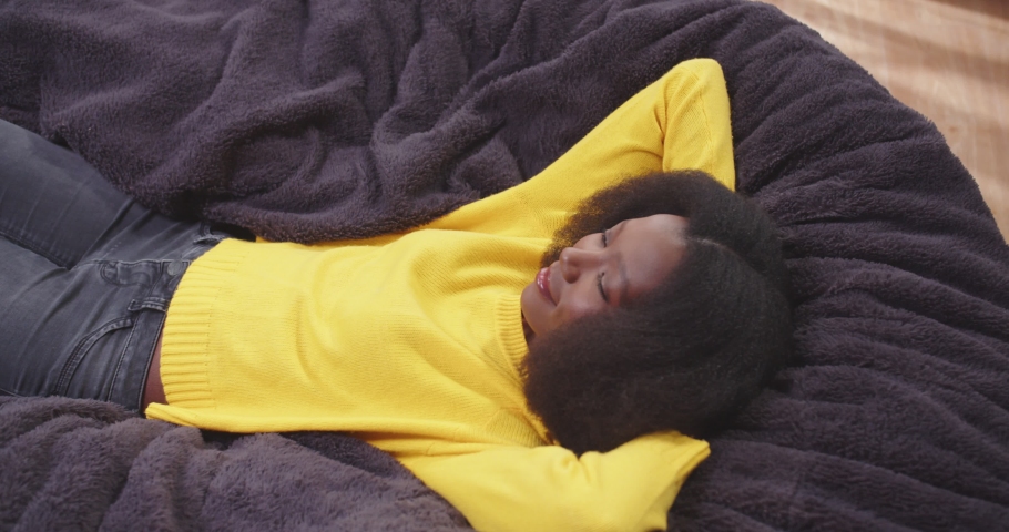 Top view of african american wooman lying on sofa with arms crossed behing her head looking around. Female in yellow clothes. Tired female after work resting. Concept of rest, carefree.