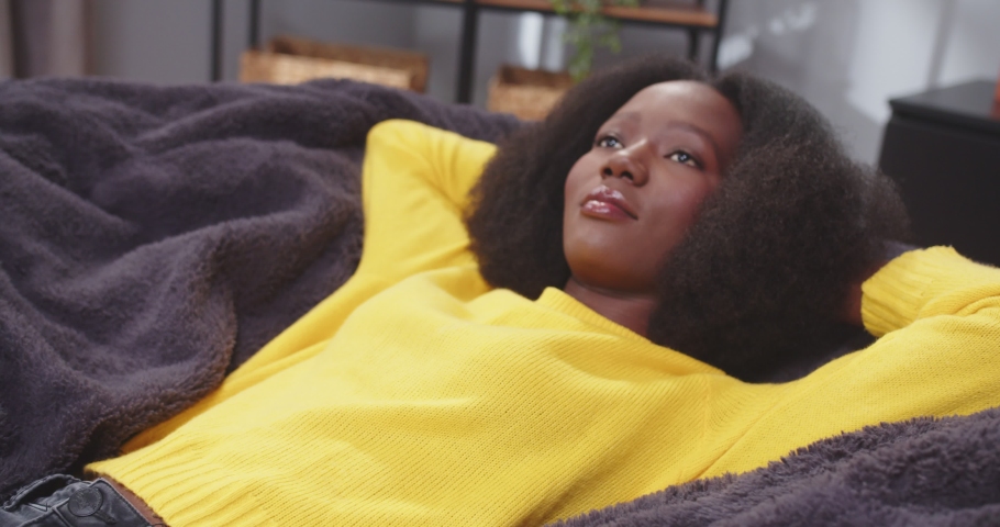 Side shot of african american wooman lying on sofa with arms crossed behing her head looking onto ceiling. Female in yellow clothes. Tired female after work resting. Concept of rest, carefree.