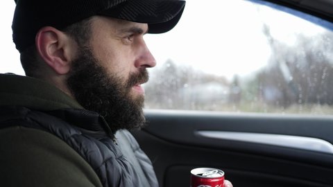 WROCLAW, POLAND - FEB 22, 2022: Handsome adult bearded man driving a car hold drinking a coca cola can sugary carbonated beverage
