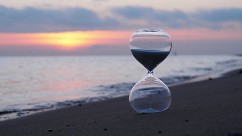Hourglass standing on the shore, small waves and a sunset sky in background, blurred backdrop. The subdued natural light, warm yellow glow of sunset through clouds, and blue hue of surrounding colors