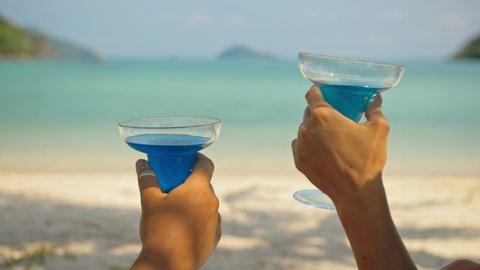 The love couple is holding a glass of blue curacao cocktail, on sea. Close up. Man and woman drink alcohol on sand beach in shadow by azure ocean of a tropical island. Celebrate summer holiday.