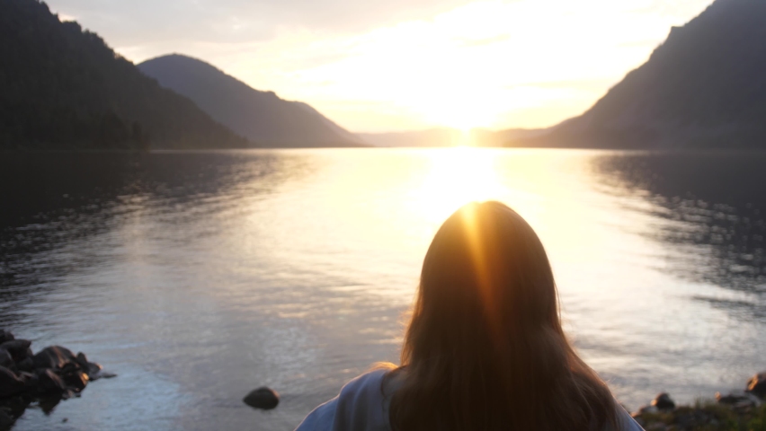 Woman raising hands and makes heart on the edge of the mountain cliff on lake at sunset