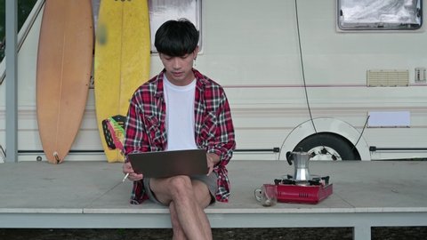 Young asian man working with laptop and drinking hot coffee at camper van on weekend. Freelancer wearing scott shirt relaxing on patio at morning