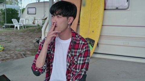 Young asian man wearing scott shirt relaxing with smoking on patio at camper van on vacation