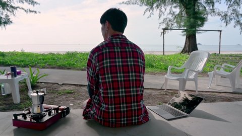 Young asian man wearing scott shirt brewing coffee and laptop at patio on the beach 
