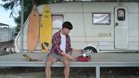 Young asian man pouring hot coffee with drinking at camper van on weekend. Freelancer wearing scott shirt relaxing on patio at morning