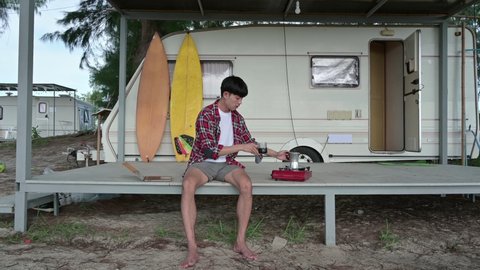 Young asian man drinking hot coffee and gas stove at camper van on weekend. Freelancer wearing scott shirt relaxing on patio at morning