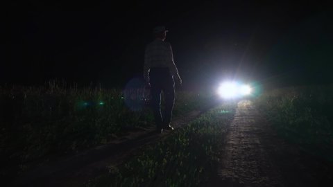 A man with a basket and a flashlight walks at night under the headlights of a car on a field dirt road.The silhouette of a man walks at midnight in the dark in a meadow in a field.Alone nightlife man