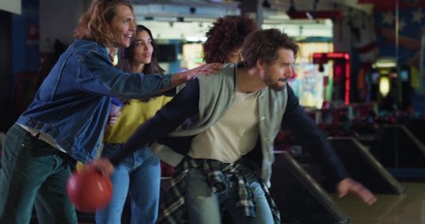 Cinematic shot of happy young male bowler throwing strike ball and exulting victory while having fun to play bowling together with multiracial college friends on weekend in sporting club.